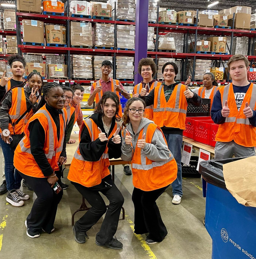 A group of students in a warehouse wearing orange vests and smiling